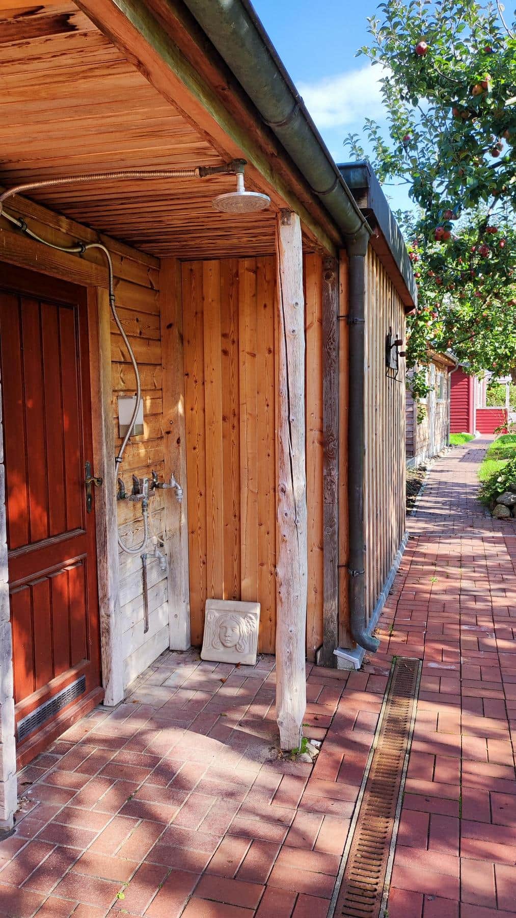 A rain shower head under a wooden canopy, with taps on the boarded wall beside it and a sandstone relief on the ground.