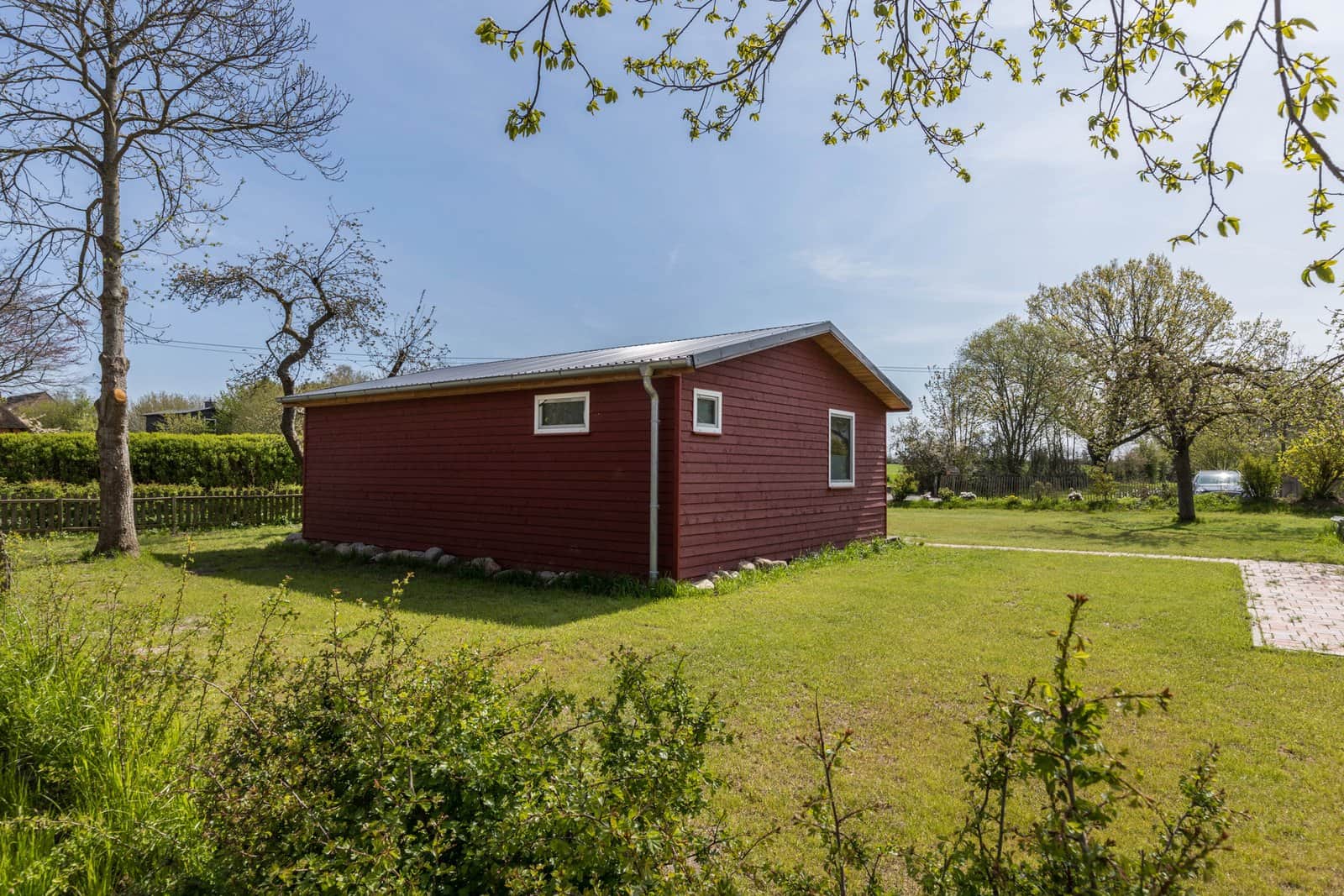 The garden from the back: a wide lawn with old fruit trees, the red timber house at its edge and hedges and open sky beyond.