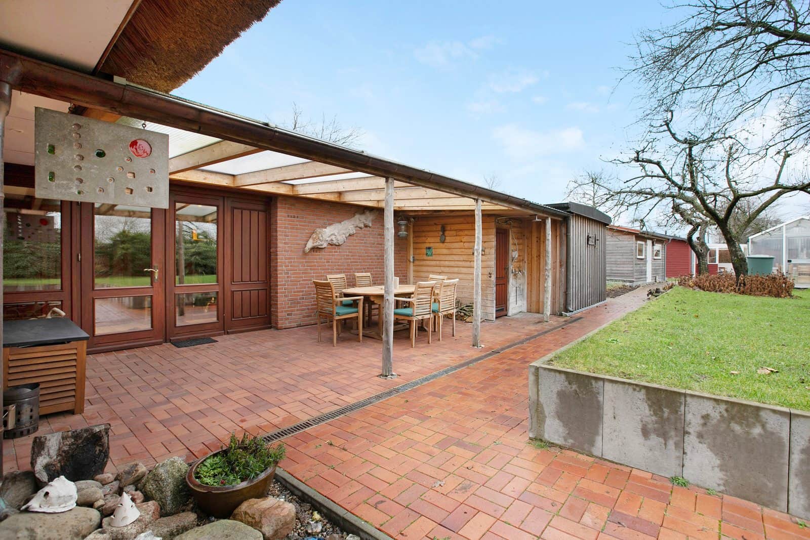 The brick path running along Olendeel under a canopy with a table and chairs; the red timber house and the greenhouse lie beyond, at the edge of the lawn.
