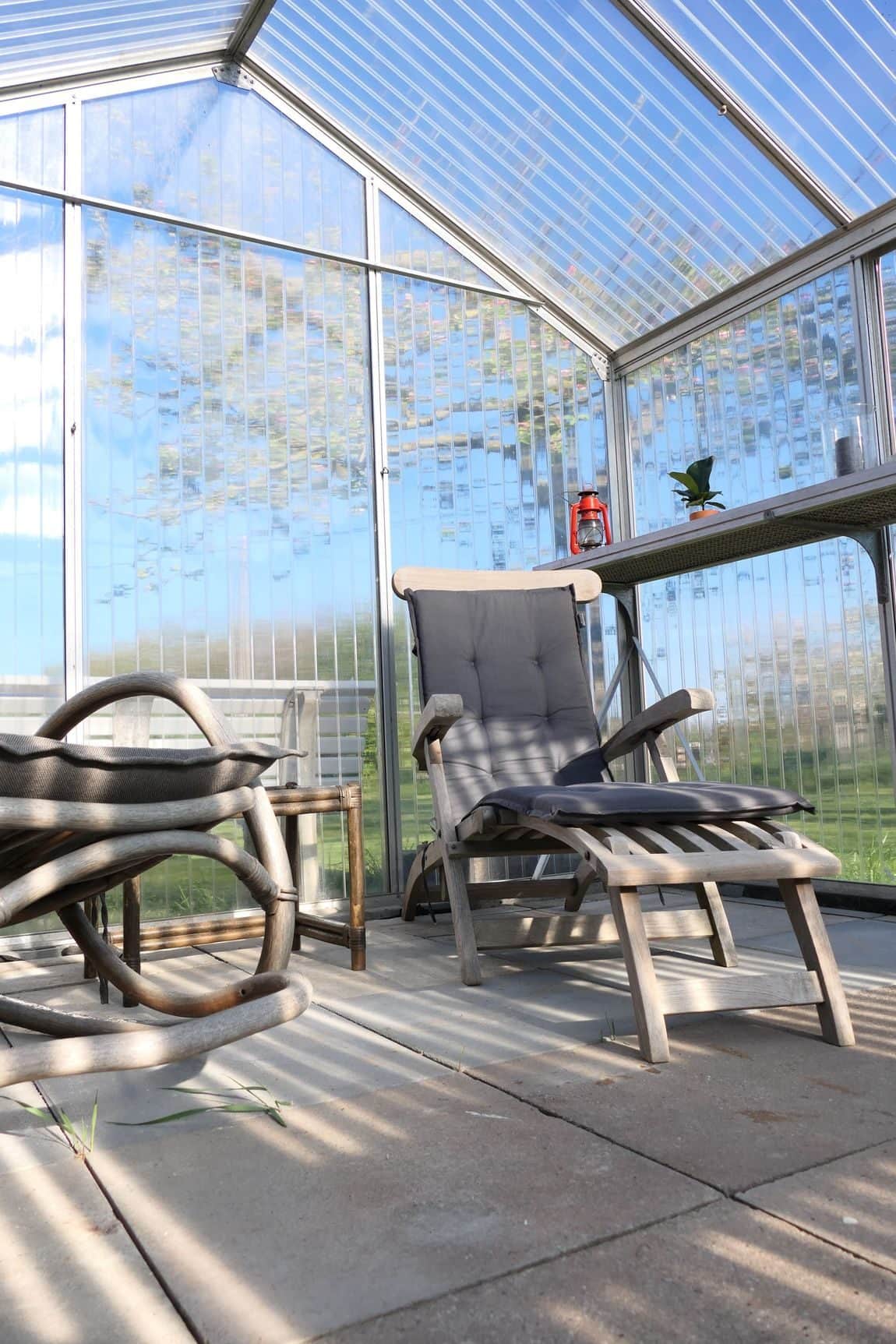 A reclining chair with a cushion, footrest extended, inside the greenhouse, with a red lantern on the side table and sun through the twin-wall panels.