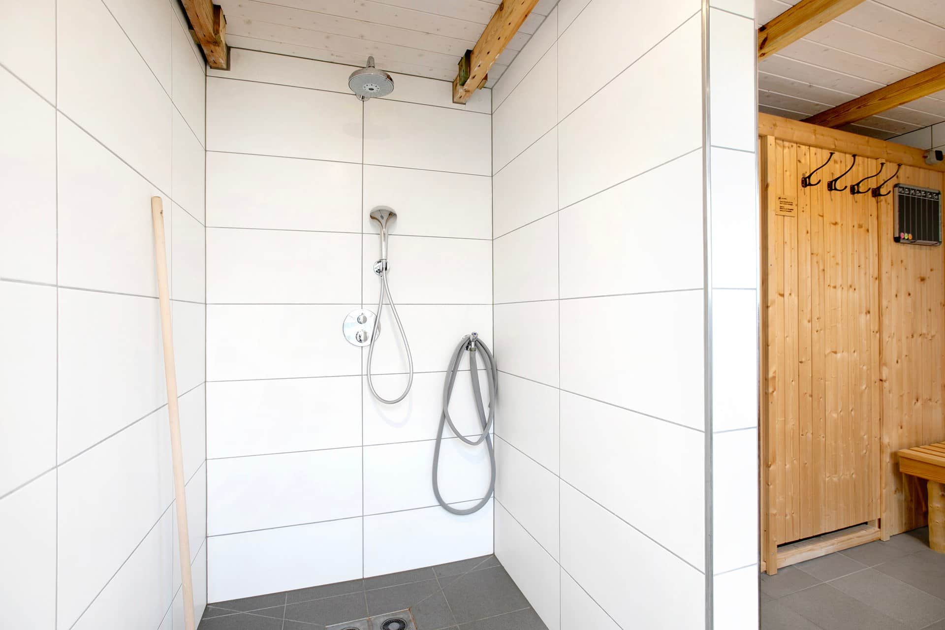 A white-tiled shower area with a rain head and hand shower, with the wooden wall of the sauna cabin and the coat hooks beside it.