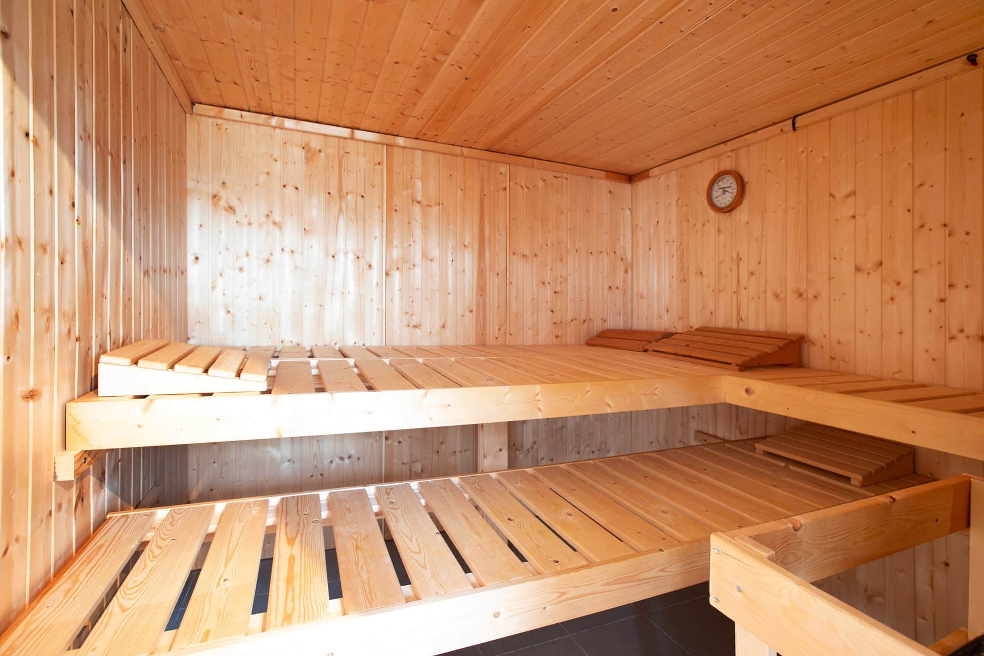 The sauna cabin in pale wood with two levels of benches around the corner, a clock on the wall and slatted wooden headrests.