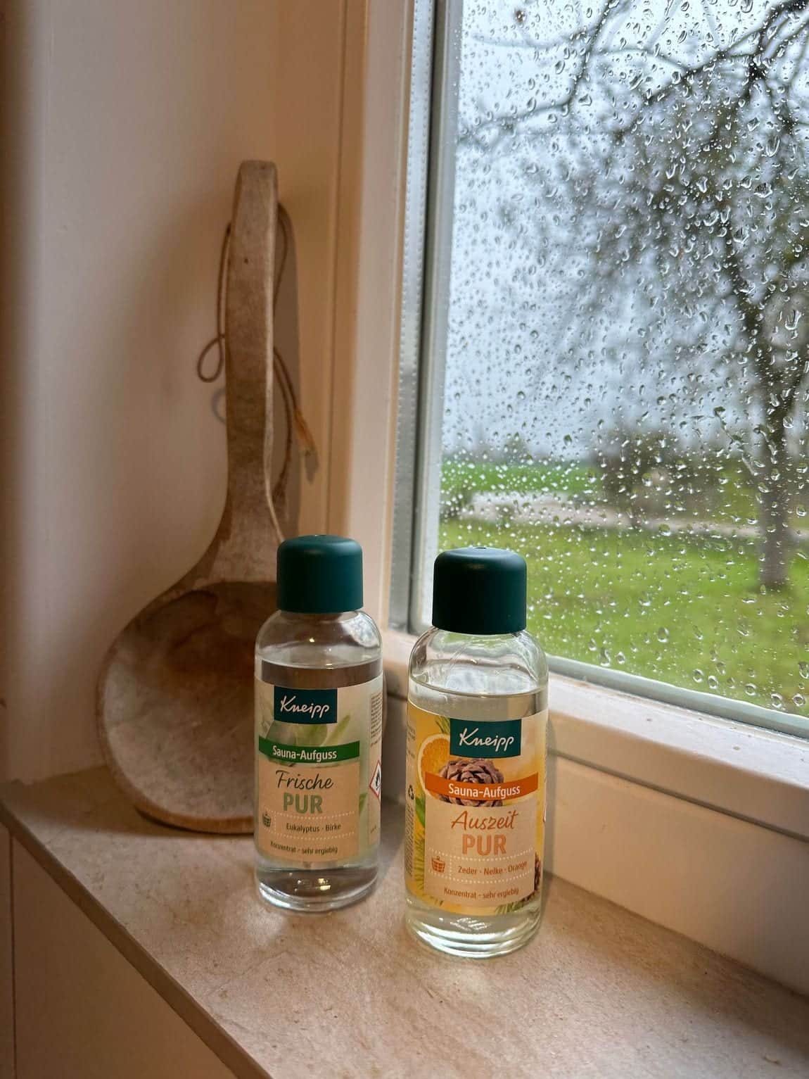 Two bottles of Kneipp sauna infusion and a wooden sauna ladle on the anteroom windowsill; outside, behind the rain-spotted pane, the meadow and a fruit tree.