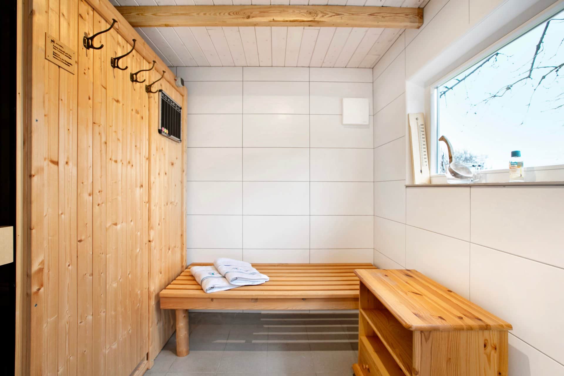The sauna anteroom: a wooden wall with four hooks above the bench, folded towels on it and a window with a thermometer to the right.