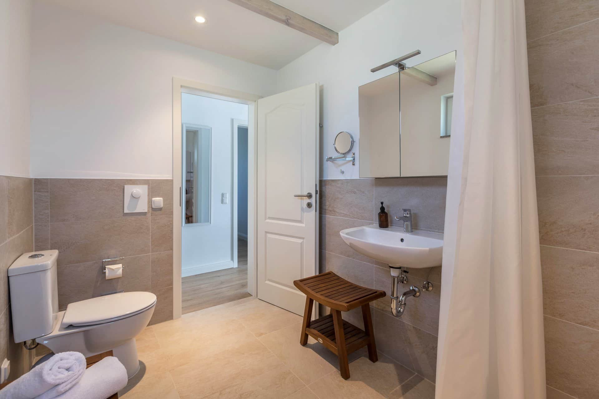 The bathroom seen from the shower: the WC on the left with a teak stool in front, the basin on the right and the open door giving onto the hallway.