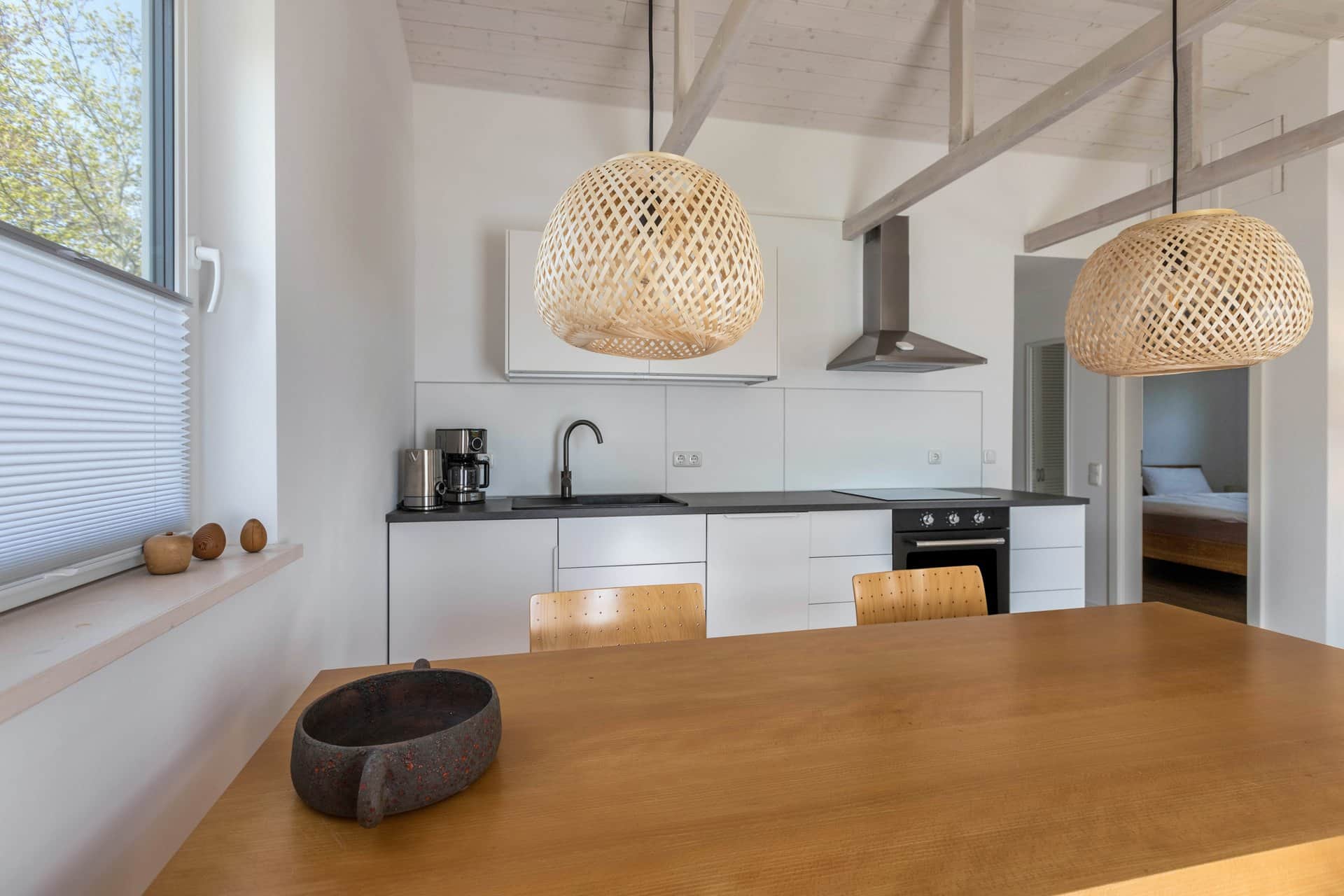 A pale wooden table under two woven pendant lamps with a stone bowl on it, and the white kitchen units with a coffee machine and oven behind.