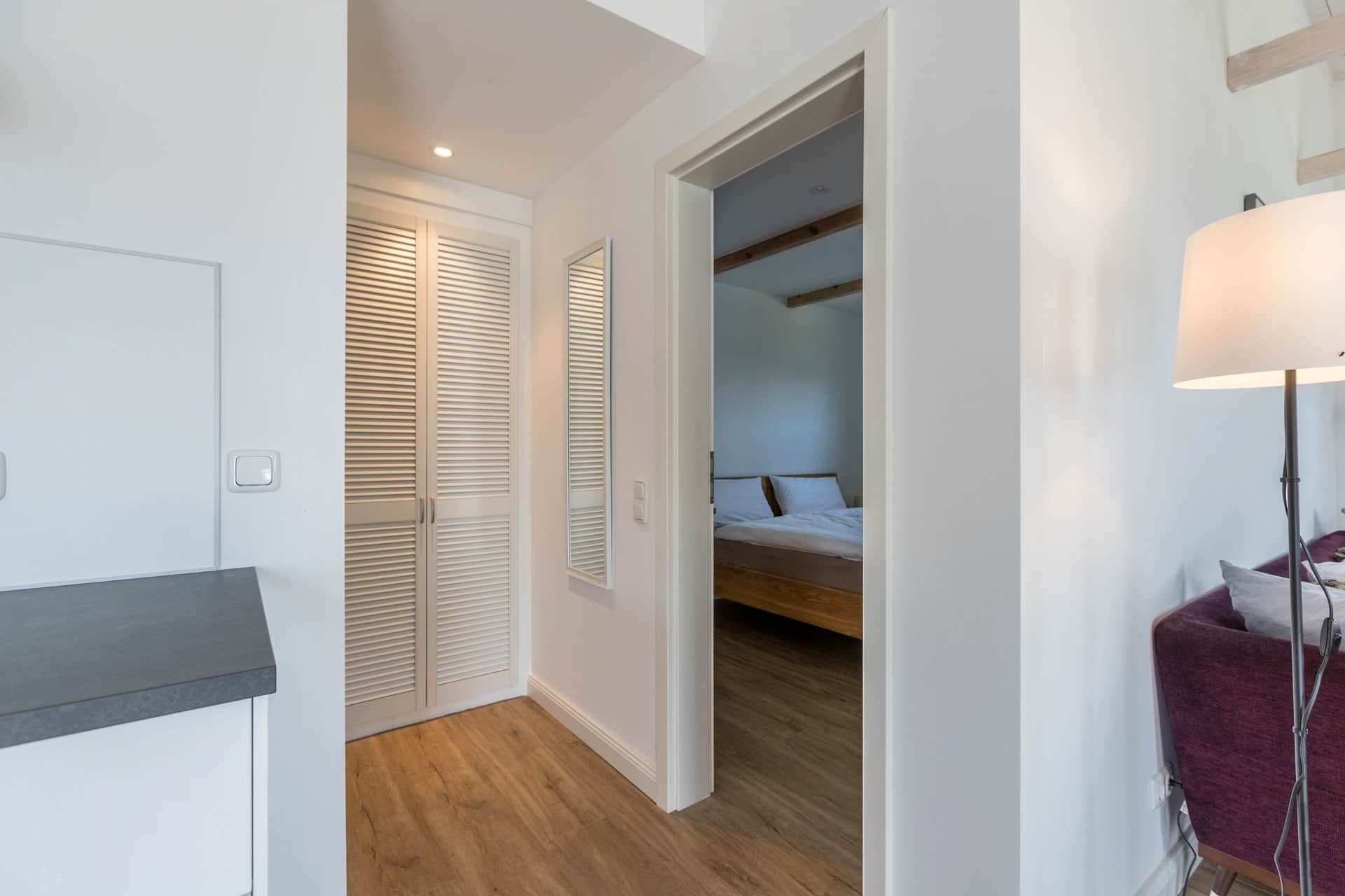 The small hallway between living room and bedroom: a louvred door conceals the fitted cupboard, and the bed is visible through the open door.