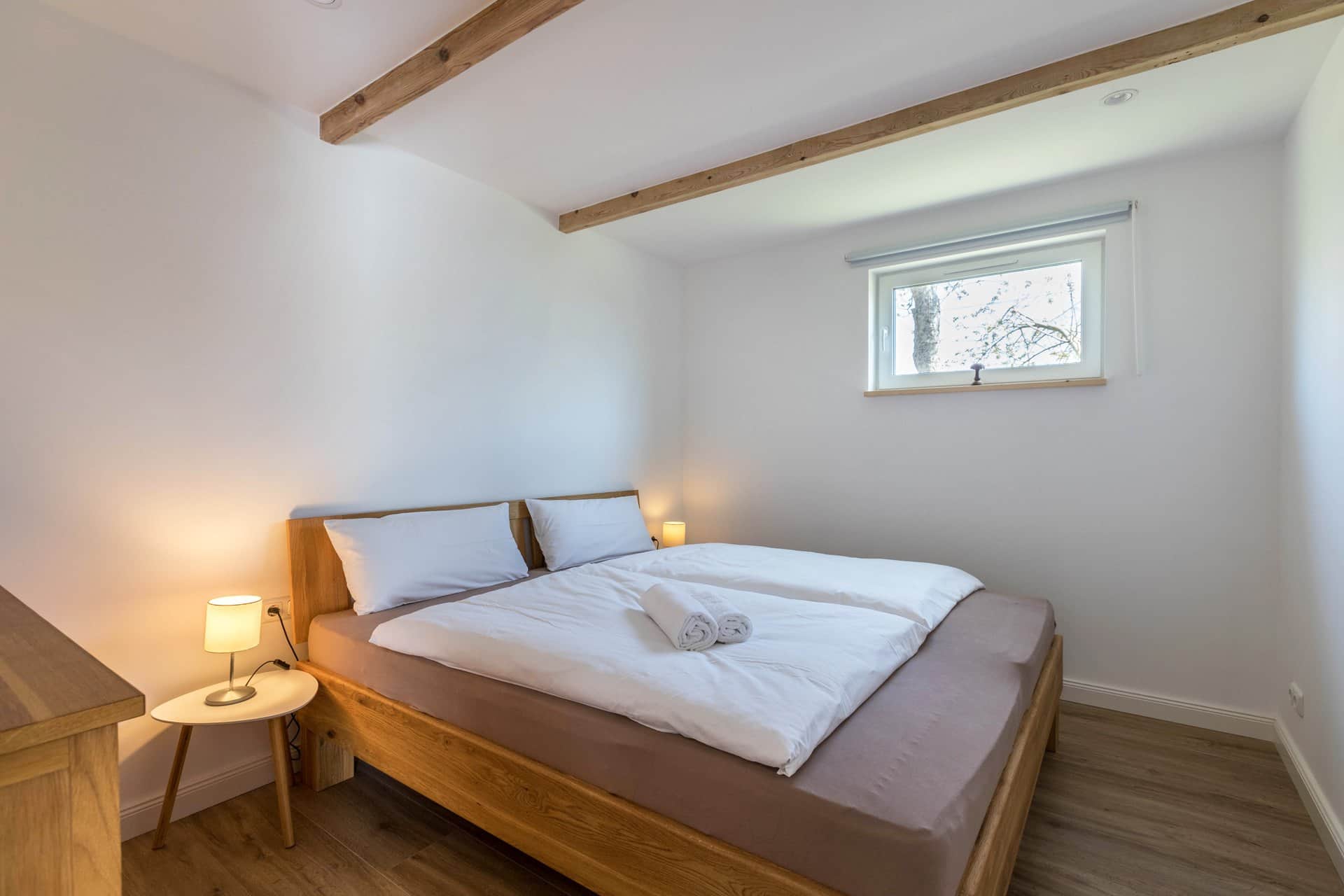 The same bedroom seen from the door: two rolled towels lie on the bed, a ceiling beam runs overhead and there is a small window to the right.