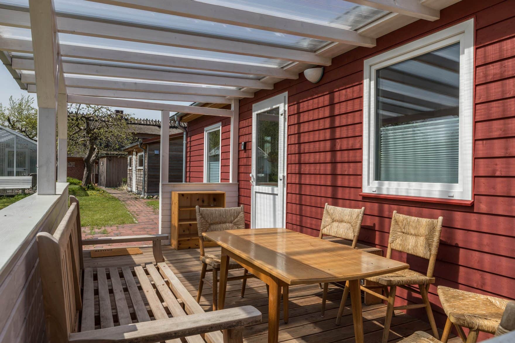 A view along the veranda: a table and four chairs against the red boarded wall, the white front door on the right and the farm outbuildings behind.