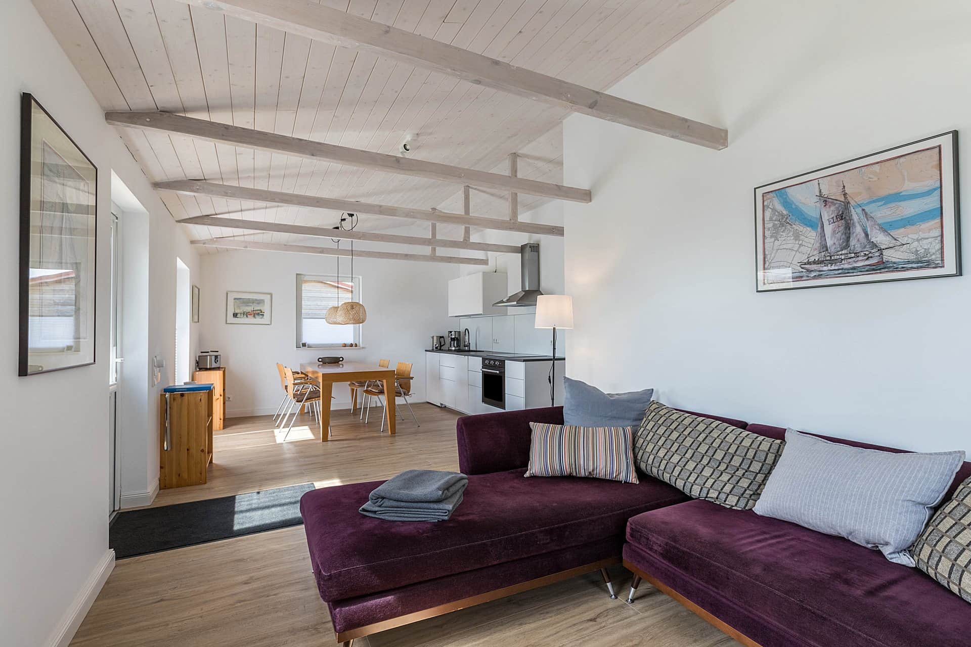 An open living room under a whitewashed beamed ceiling: the purple corner sofa in front, the dining table and kitchen units behind and a picture of a sailing boat on the wall.