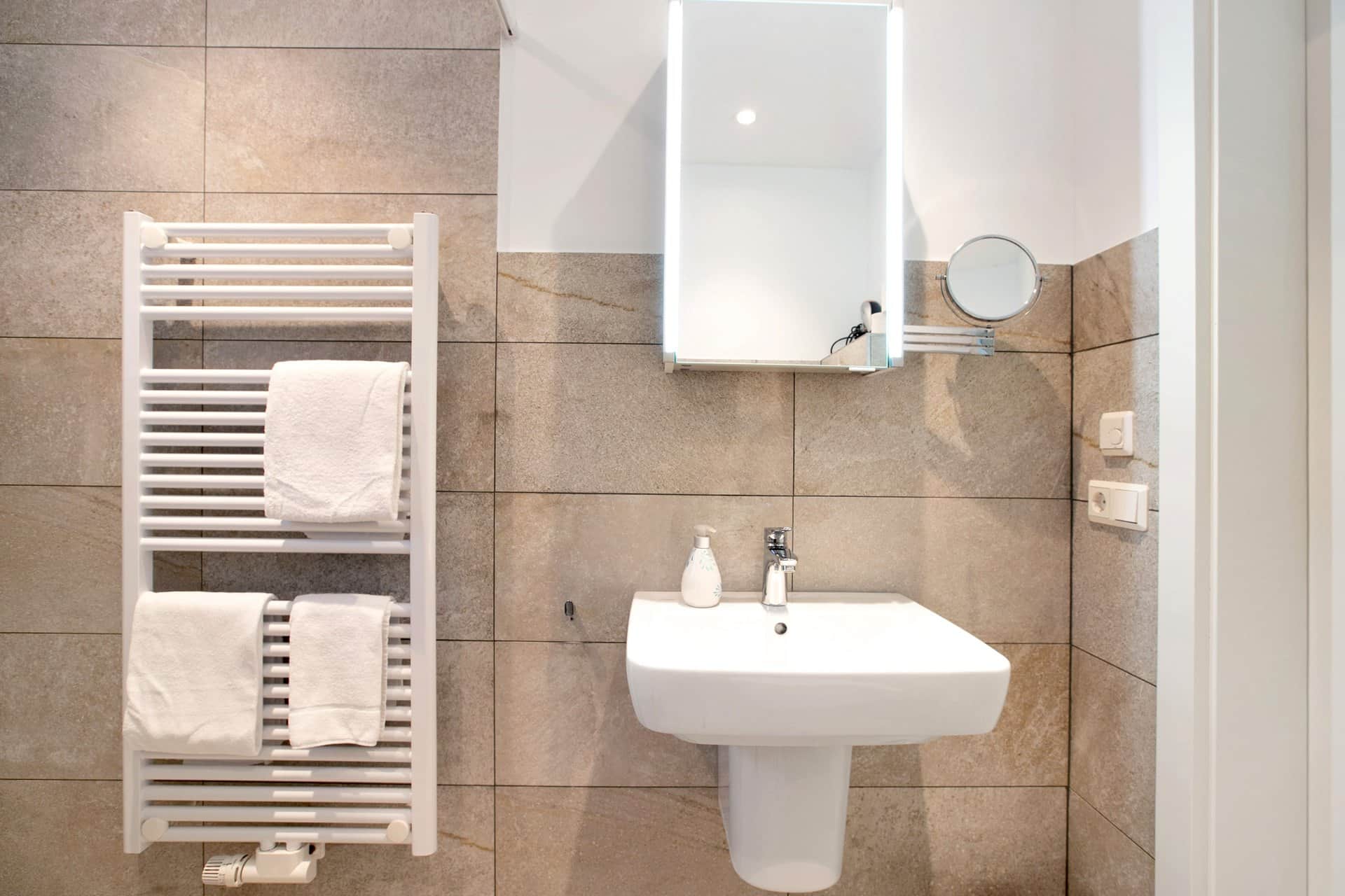 A white heated towel rail with folded towels beside the basin, with the mirrored cabinet and a shaving mirror above.