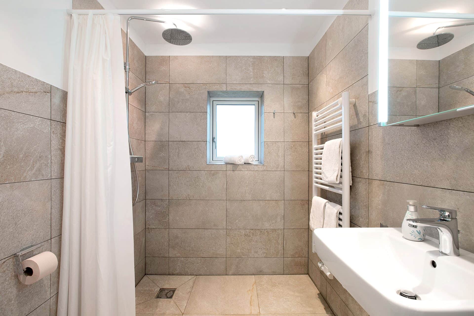 A bathroom with grey tiles: a level-access shower with a rain head behind a pale curtain and the basin under a lit mirror on the right.