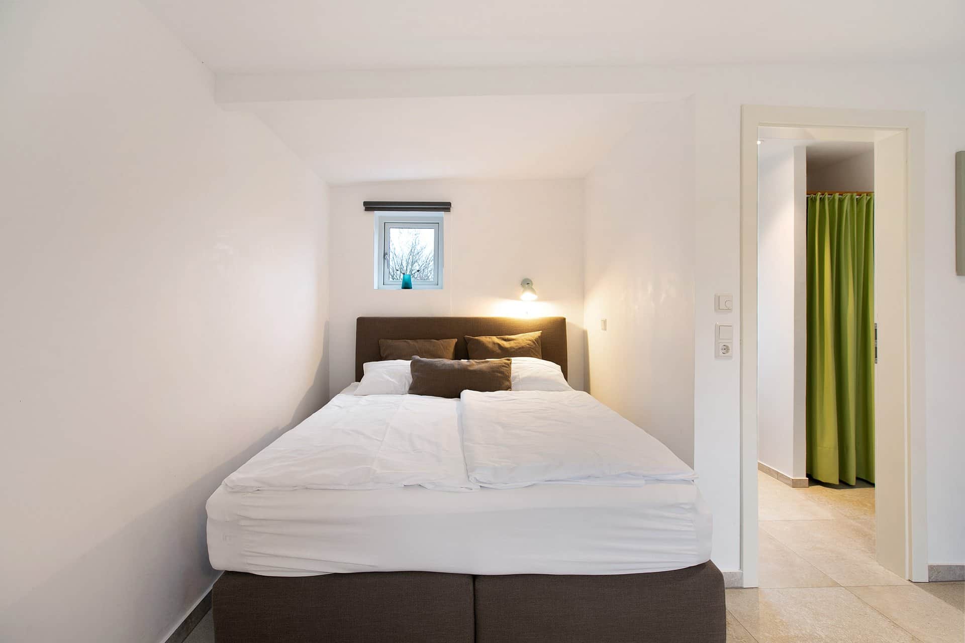 The box-spring bed in its alcove under a small window, with a green curtain and the open door to the hallway beside it.