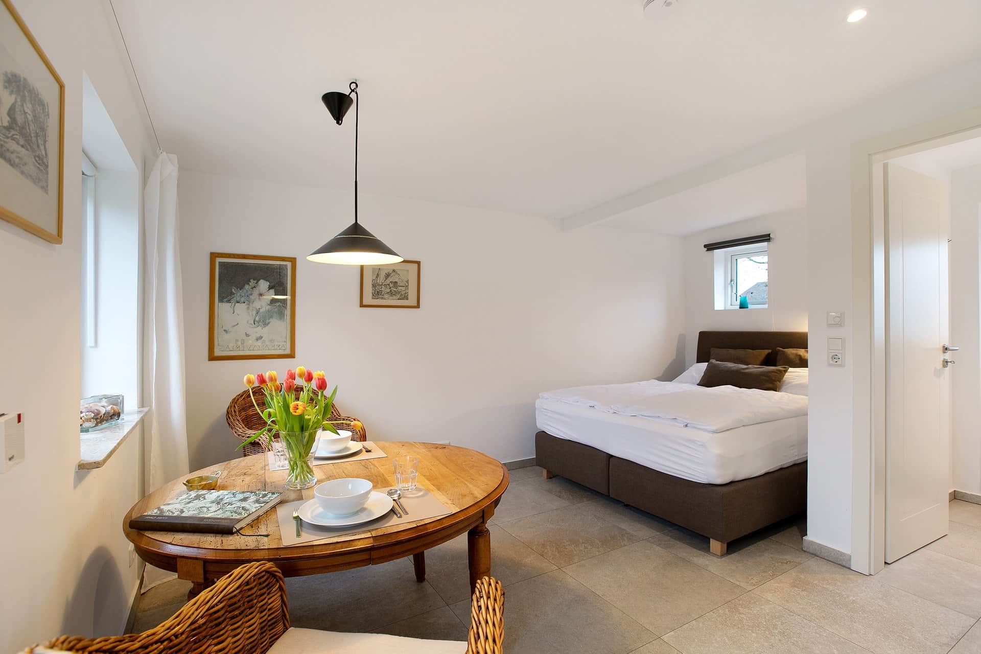 The laid table at the window with tulips and an open book, with the bed and the bathroom door behind.