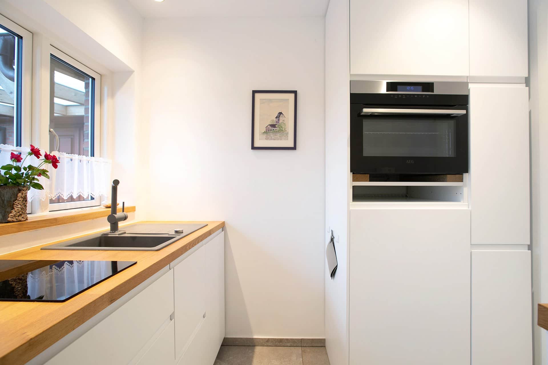 The kitchen seen from the door: the oven in a tall unit on the right, the worktop at the window on the left and a framed drawing between them.