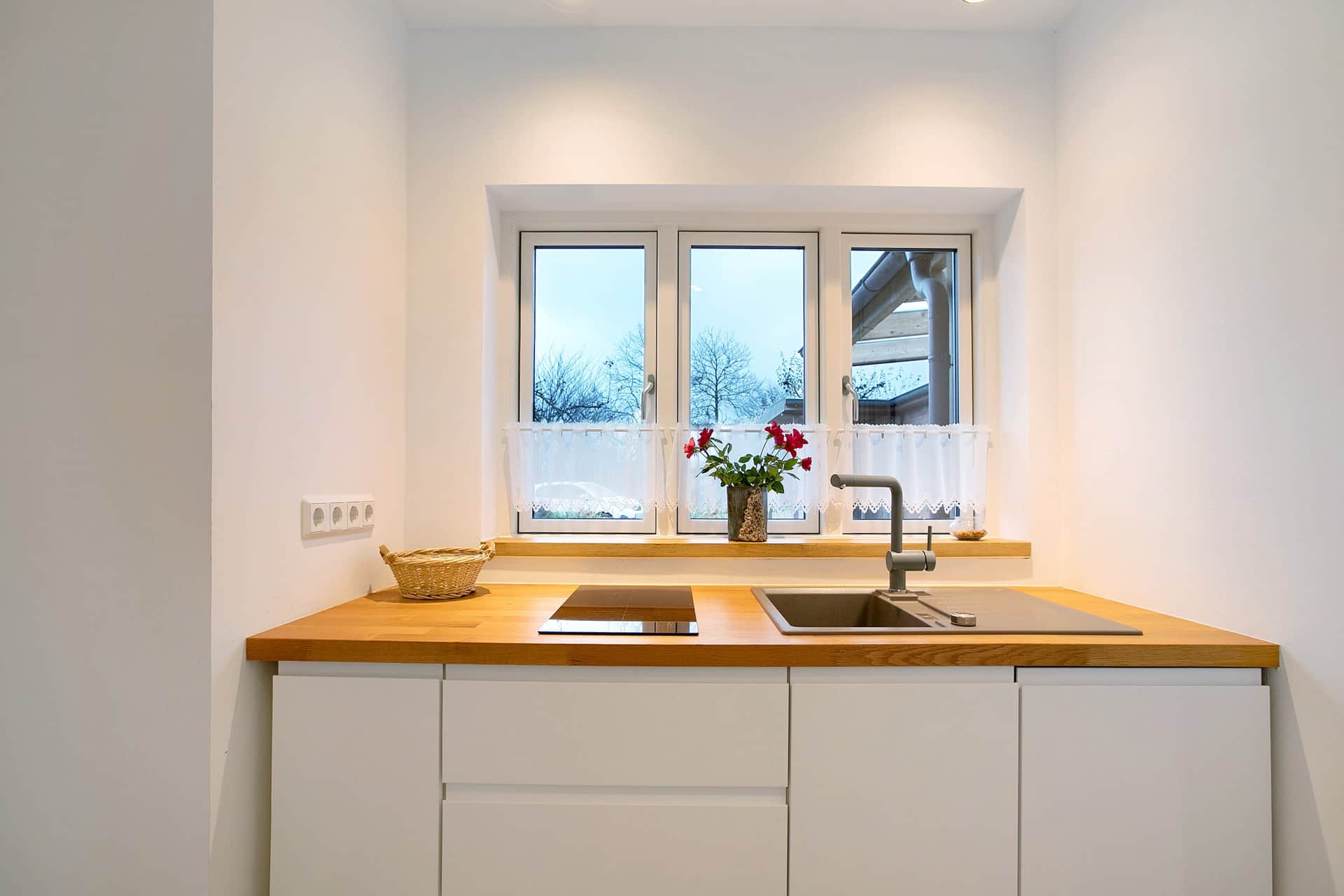 White kitchen units with a wooden worktop under the window, a basket and flowers on it, the induction hob to the left and the sink to the right.