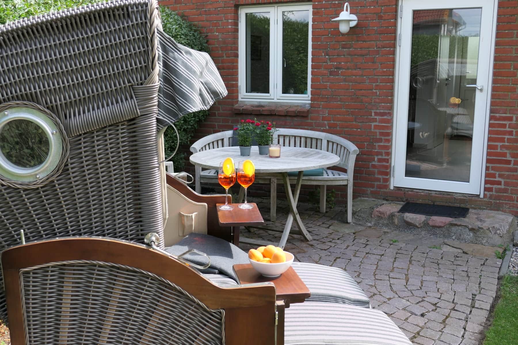 The view out of the beach chair: two aperitif glasses and a bowl of oranges on the pull-out trays, with the garden table, the bench and the Lüttdeel’s terrace door beyond.