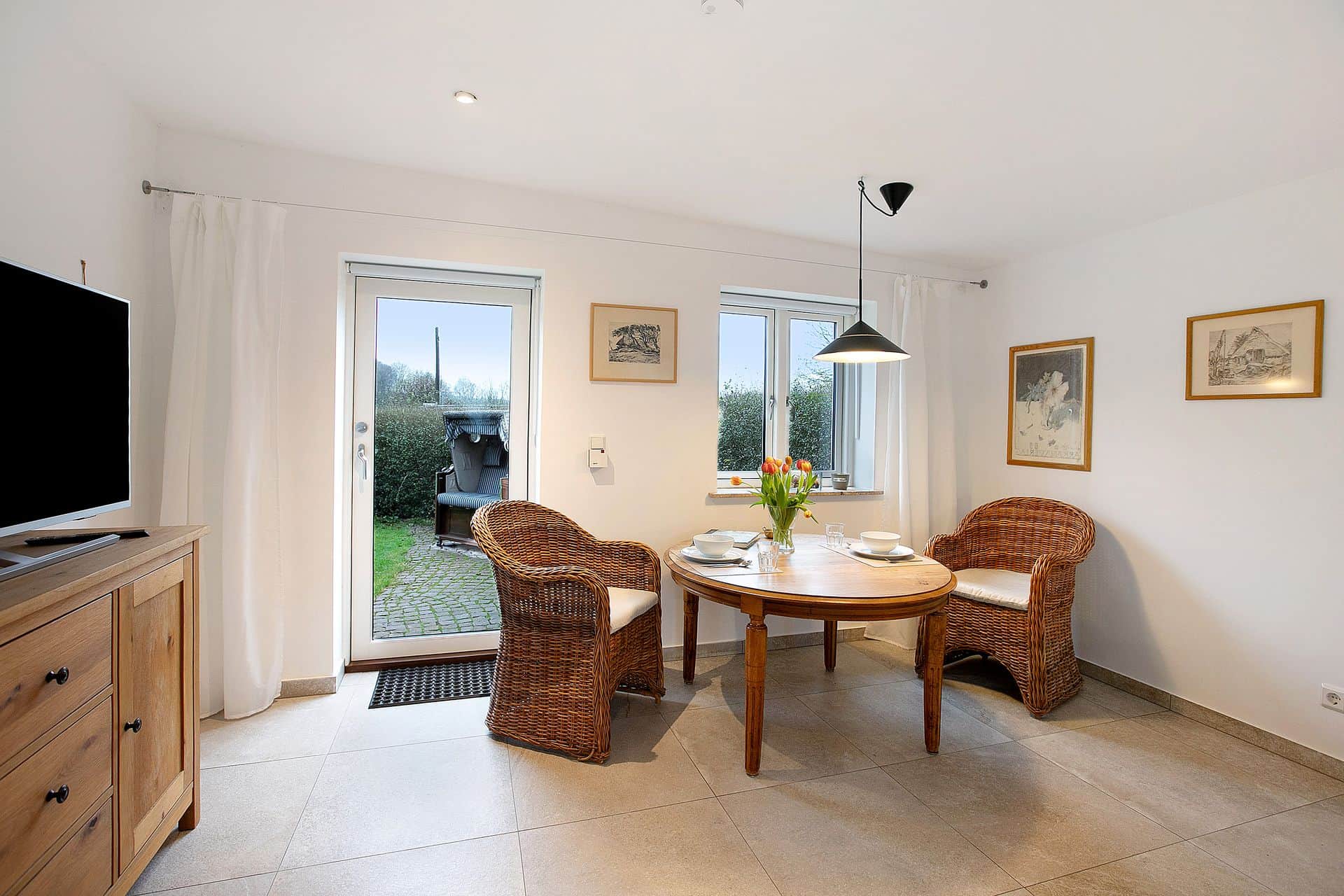 A round table with two wicker chairs by the terrace door, a chest of drawers with a television on the left and framed drawings on the wall.