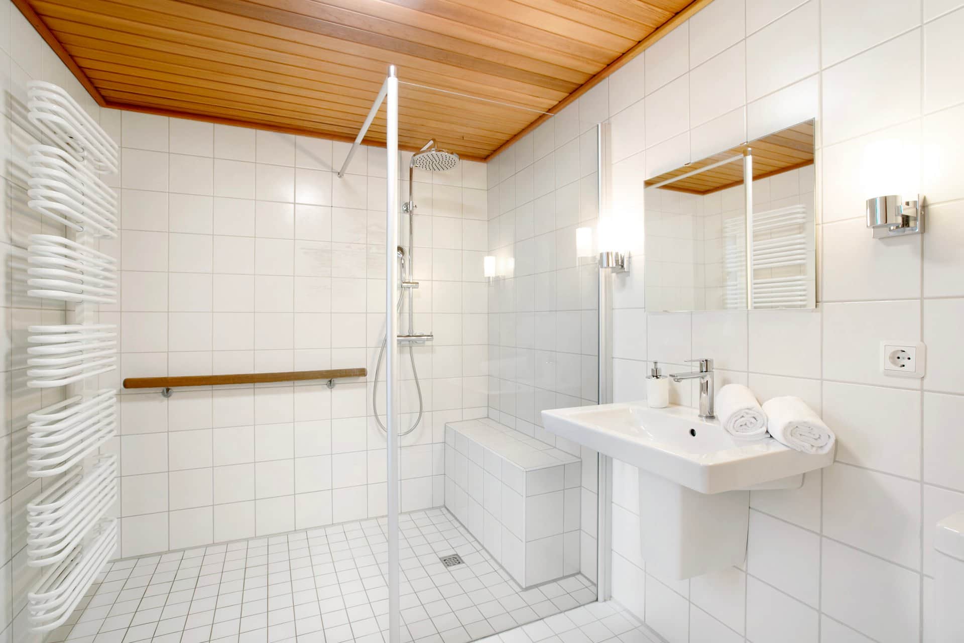 A white-tiled bathroom under a wooden ceiling: a level-access shower behind glass, a heated rail full of white towels on the left and the basin on the right.