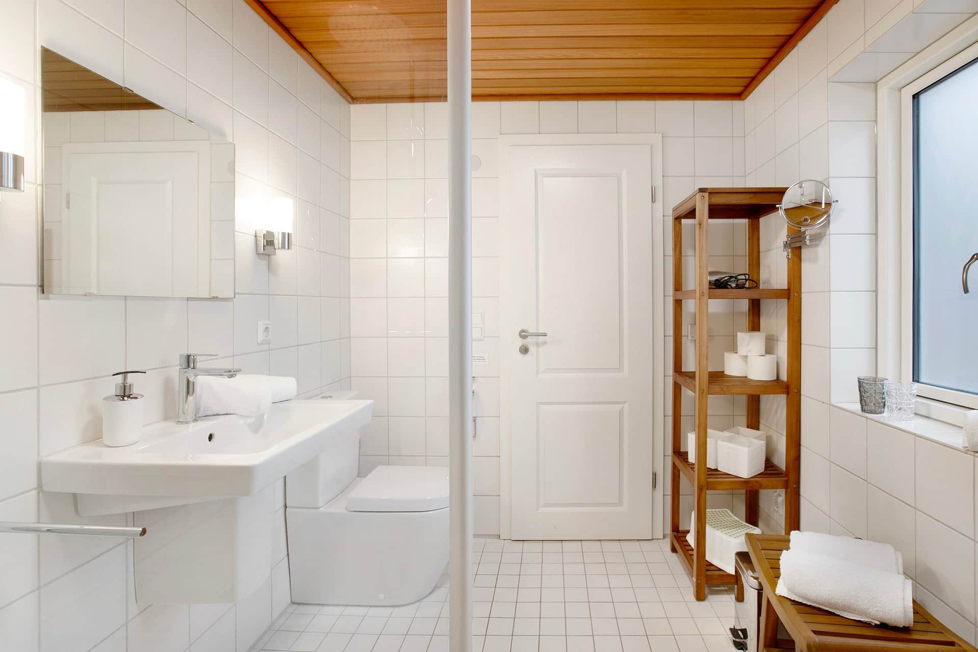 The same bathroom from the other side: the basin and WC against the tiled wall, with a wooden shelf of towels beside them.