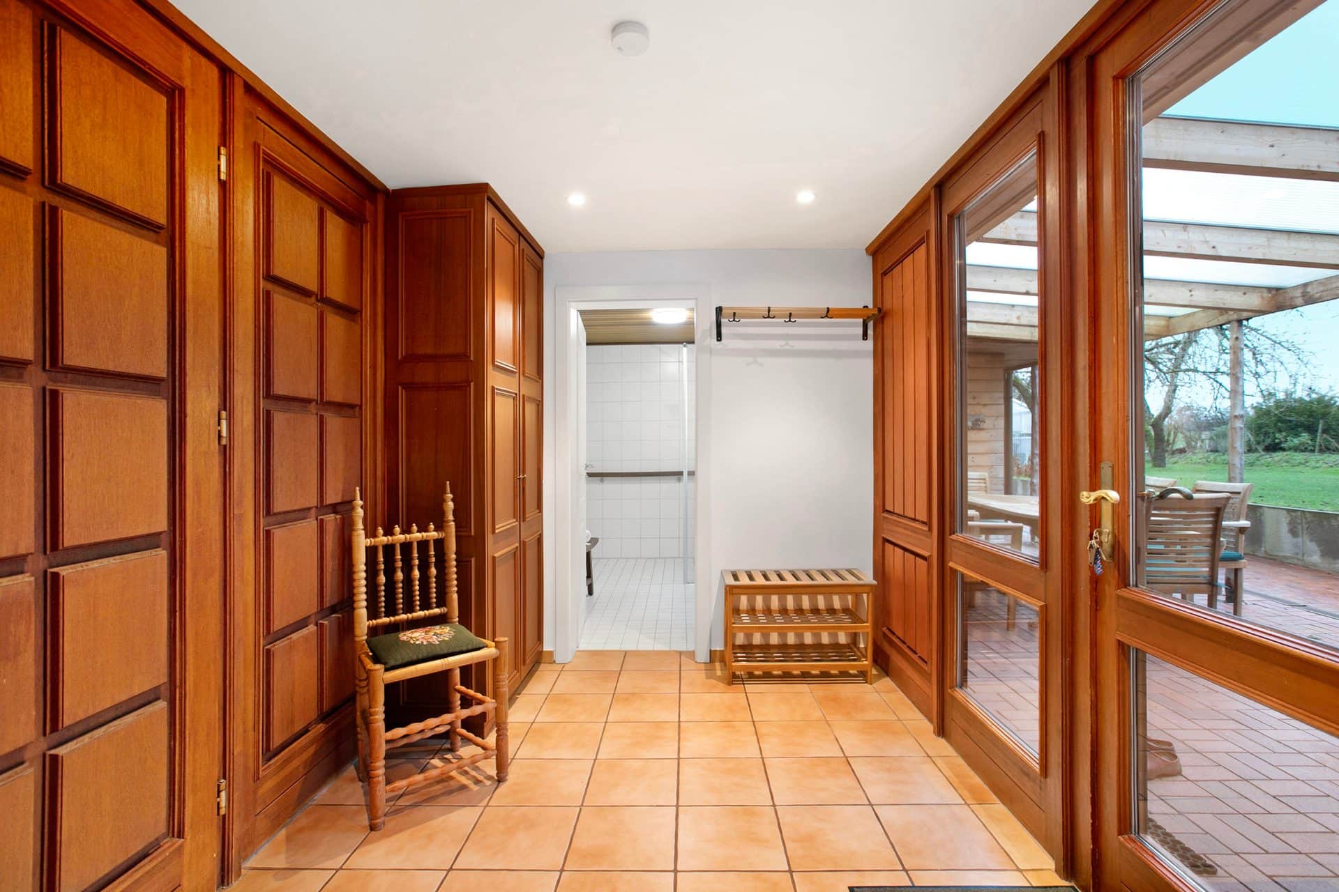 A hallway with full-height wood panelling and fitted cupboards, a tiled floor and a chair against the wall; glazed doors lead to the covered terrace on the right.