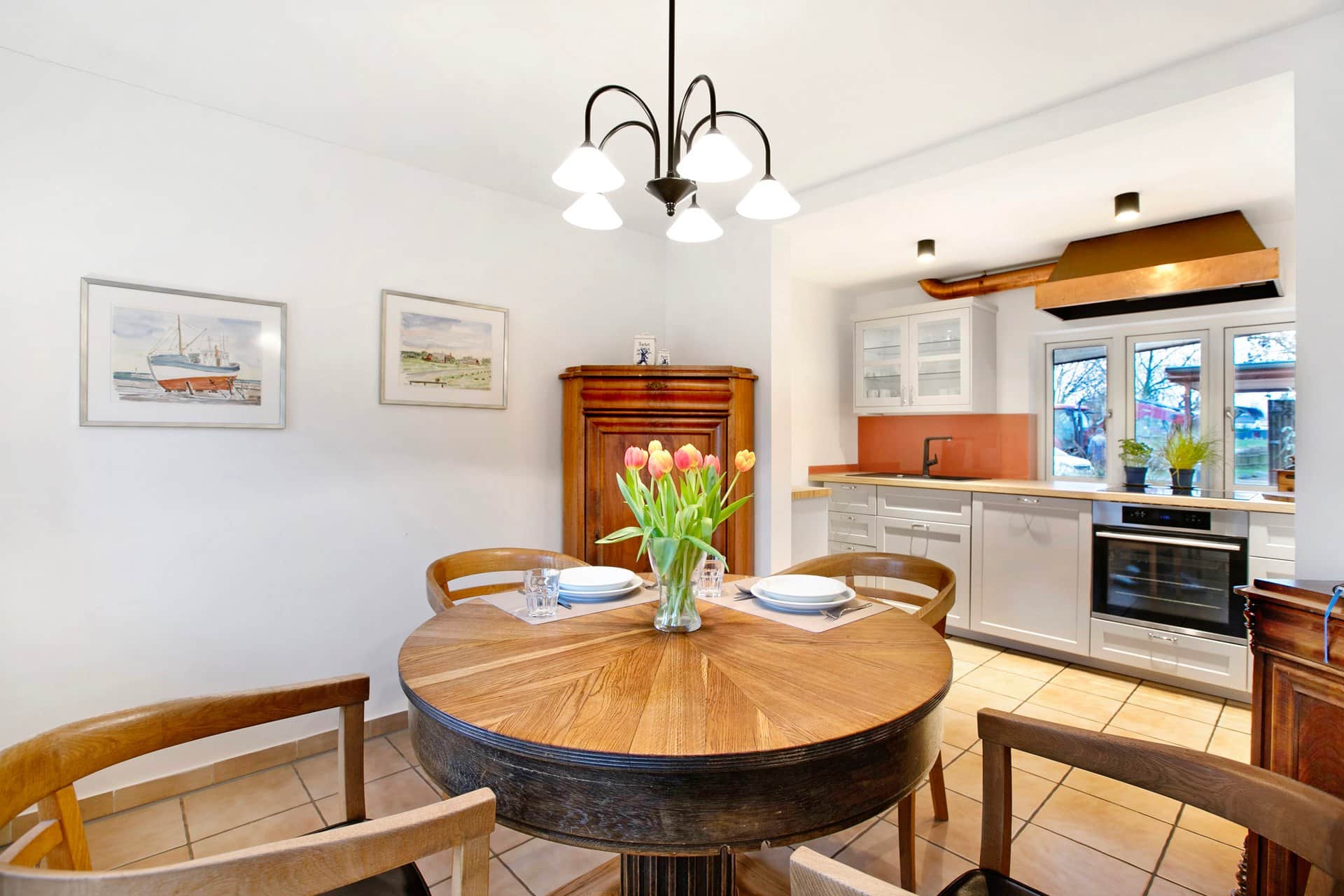 A round wooden table laid for four with tulips in the middle, a wrought-iron light above and the kitchen units with an orange splashback behind.