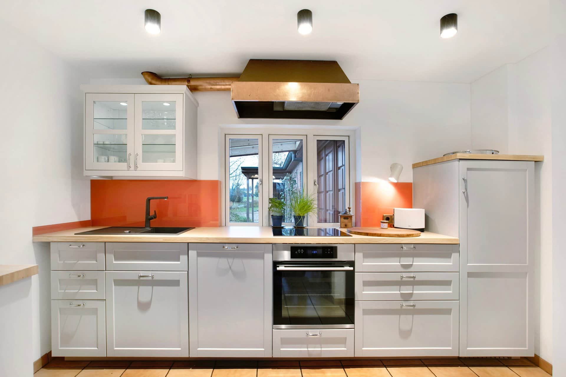 White kitchen units with a wooden worktop and an orange splashback holding a sink, oven and induction hob, with a boxed-in extractor hood above.