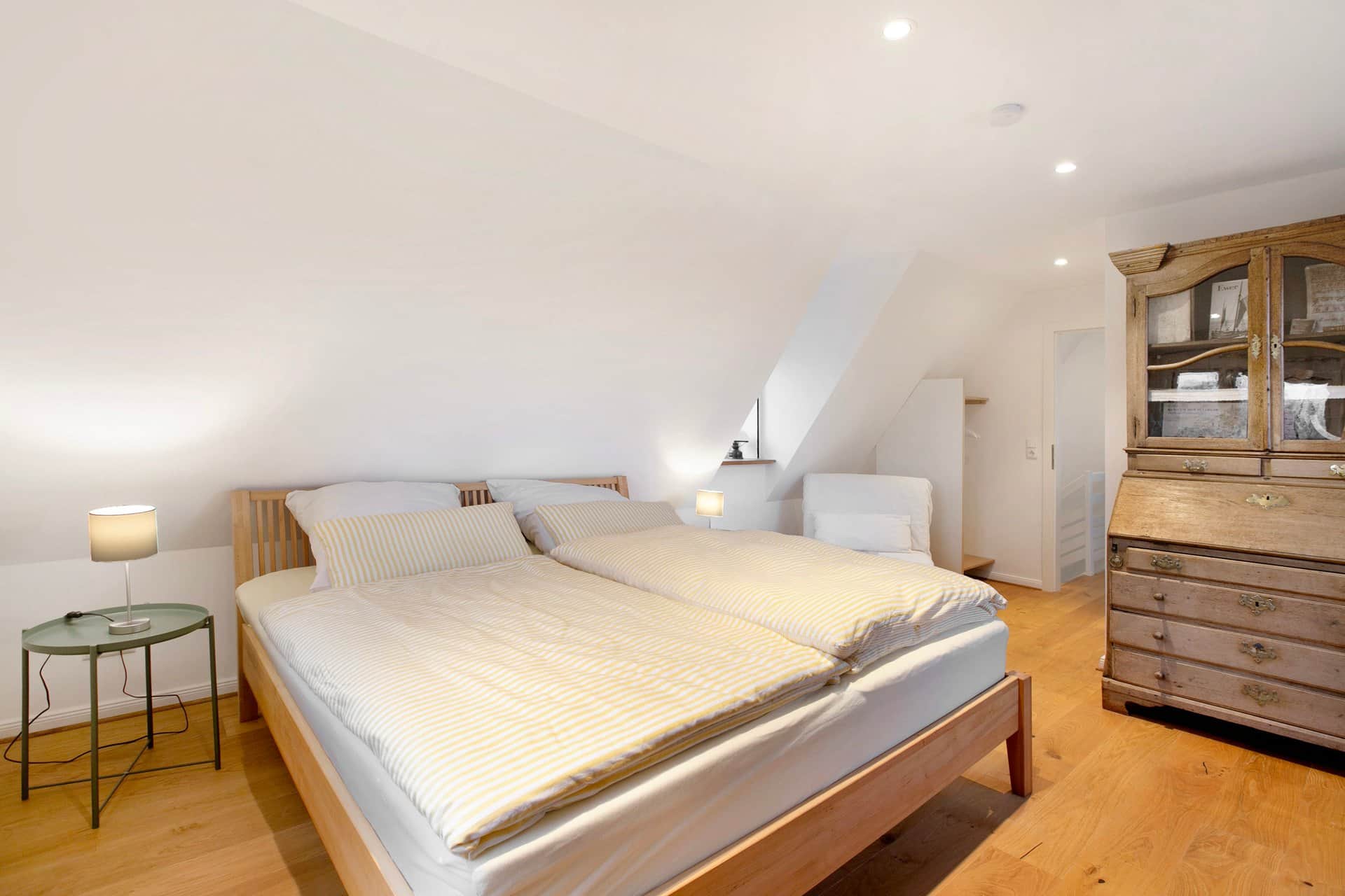 The second bedroom under the sloping roof with yellow-and-white striped bedding, two reading lamps and an antique bureau with a glazed top.