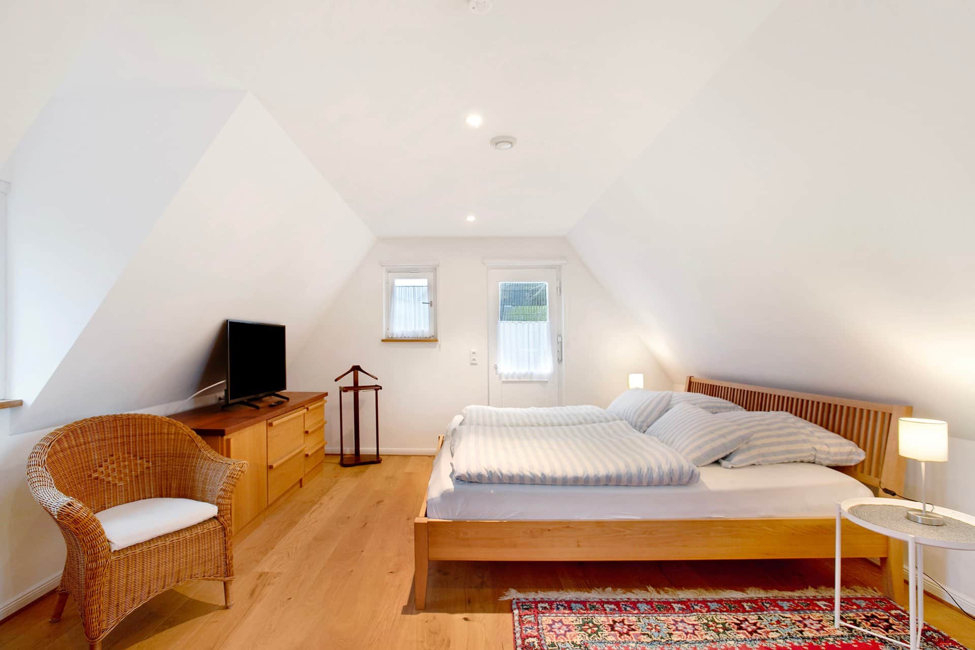 A bedroom under the sloping roof: a double bed with blue-and-white striped linen, a wicker chair in front and a chest with a television to the left.