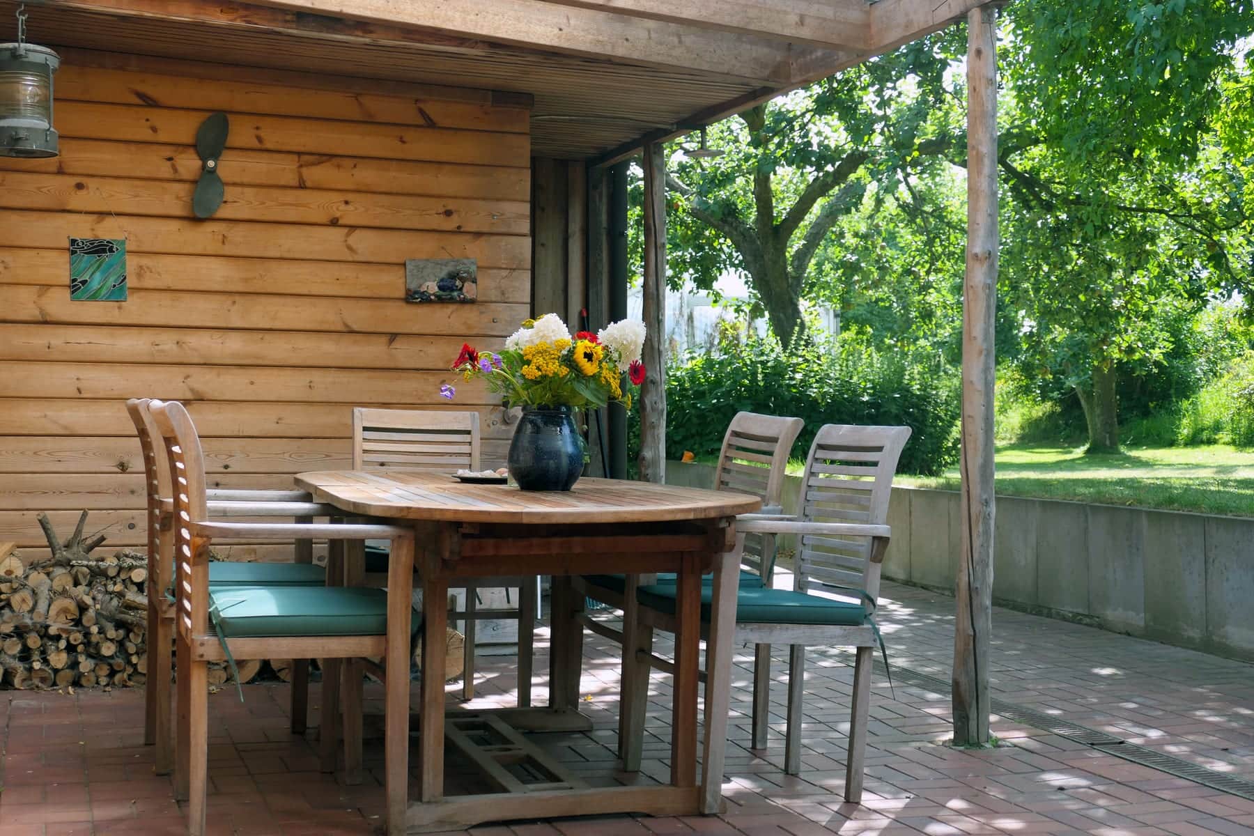 The covered terrace with an oval teak table and chairs with turquoise cushions, a bunch of flowers on the table; stacked firewood on the left and the summer garden on the right.