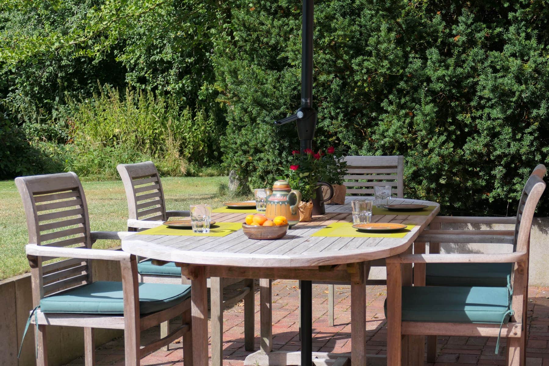 A laid teak table with six chairs on the open terrace, set with glasses, a jug and a bowl of fruit, with a tall yew hedge behind.
