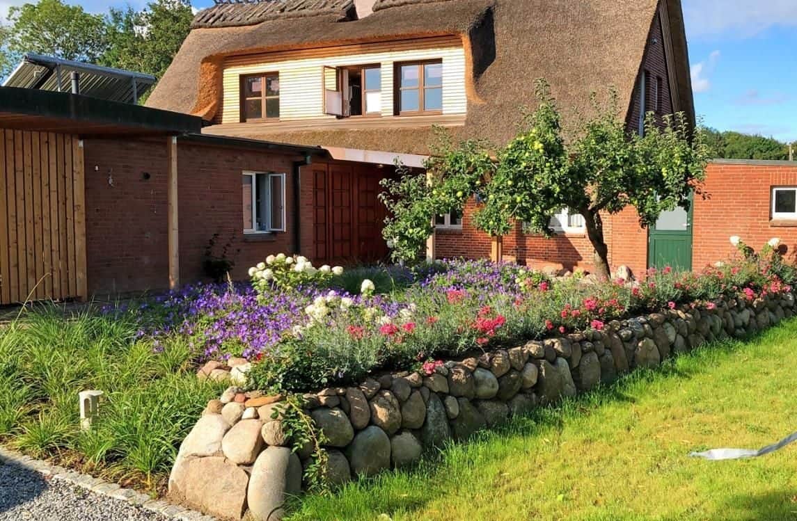 A perennial bed in flower behind a fieldstone wall, with the entrance, an apple tree and a green door behind it.