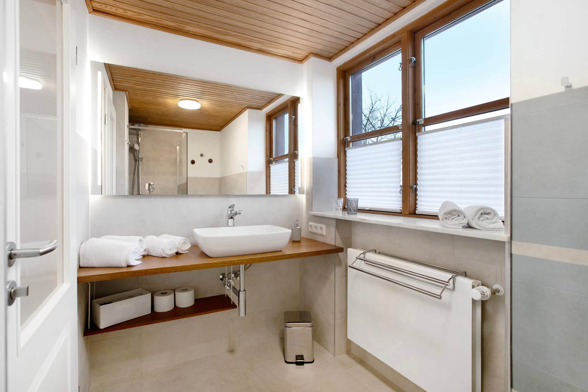 The second bathroom in grey: a countertop basin on a wooden plank in front of the mirror, with an open shelf of rolled towels beneath.