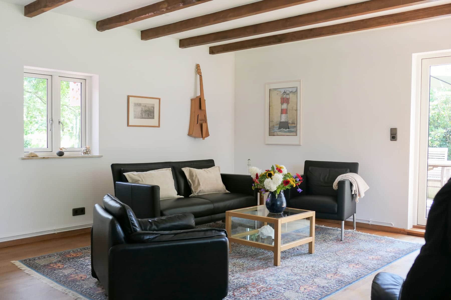 A living room under exposed ceiling beams: a black leather sofa and two armchairs around a glass coffee table with flowers, a lighthouse picture and a wooden guitar shape on the walls.