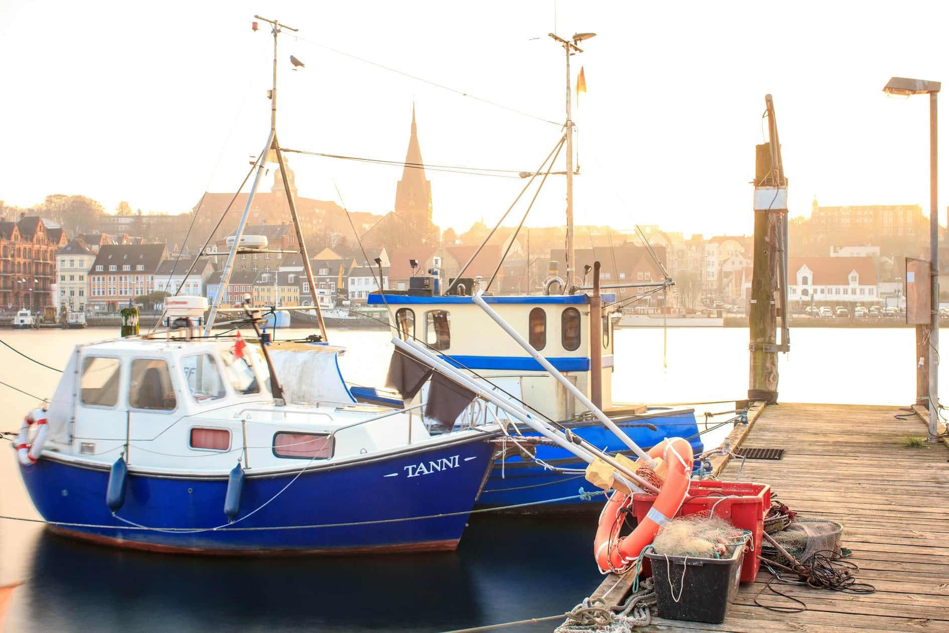 A blue-and-white fishing boat at a wooden jetty in backlight, life rings beside it and the town with a church tower behind.