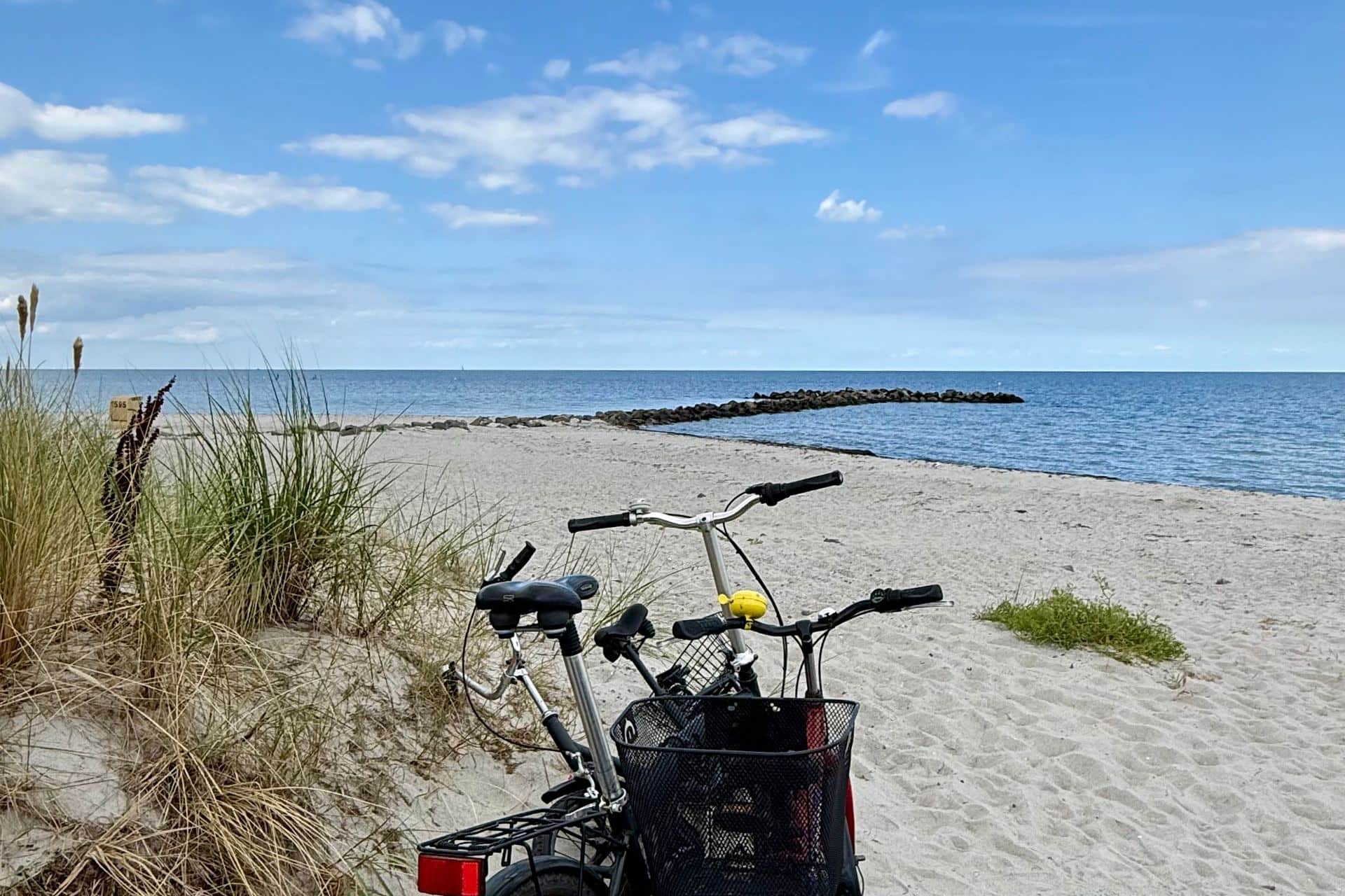 Two bicycles on the sand beside marram grass, with a stone groyne running out into the water behind.