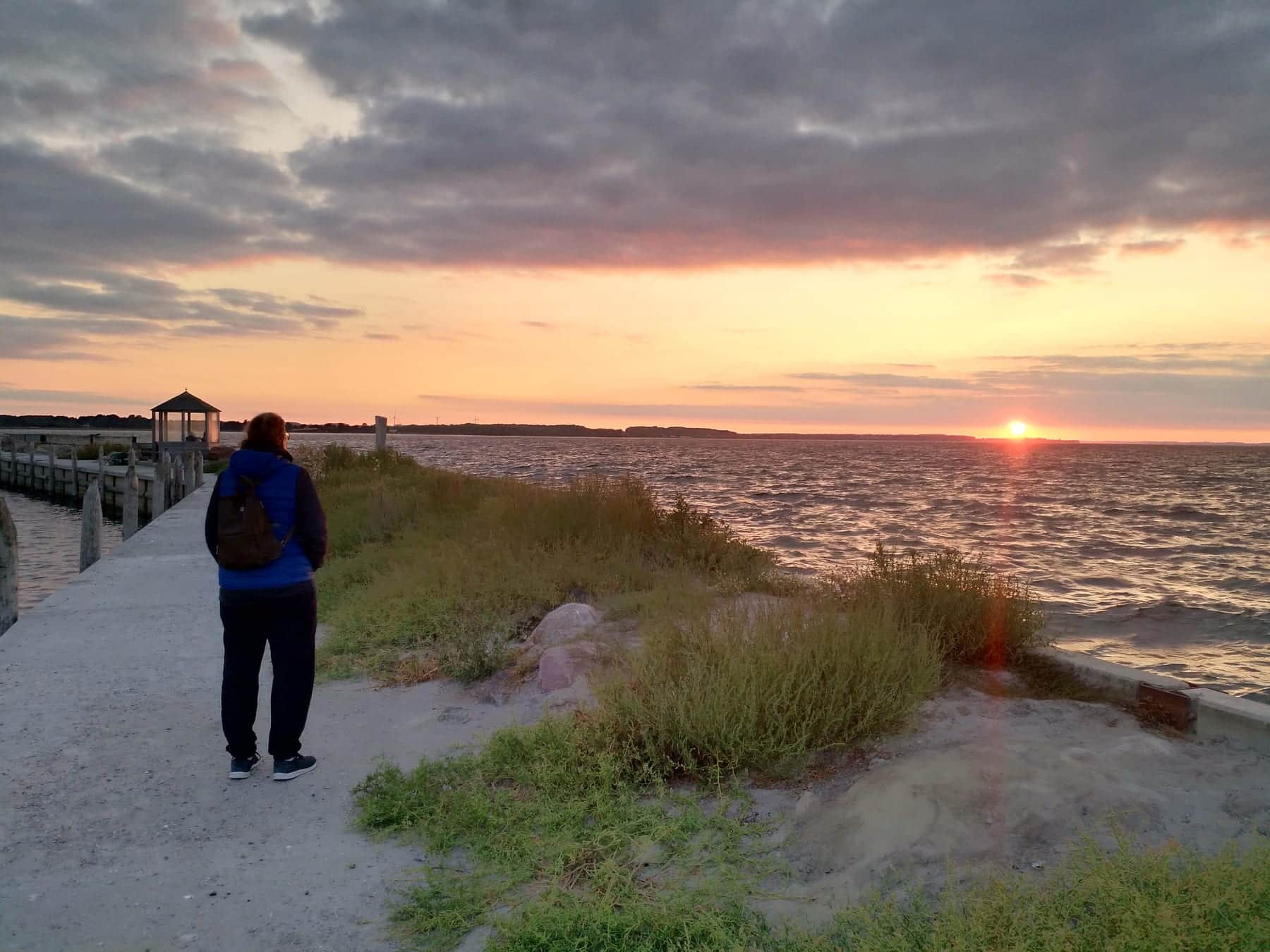 Evening on the bay: someone stands at the end of a concrete jetty looking at the sun going down over the water, a small pavilion to the left.