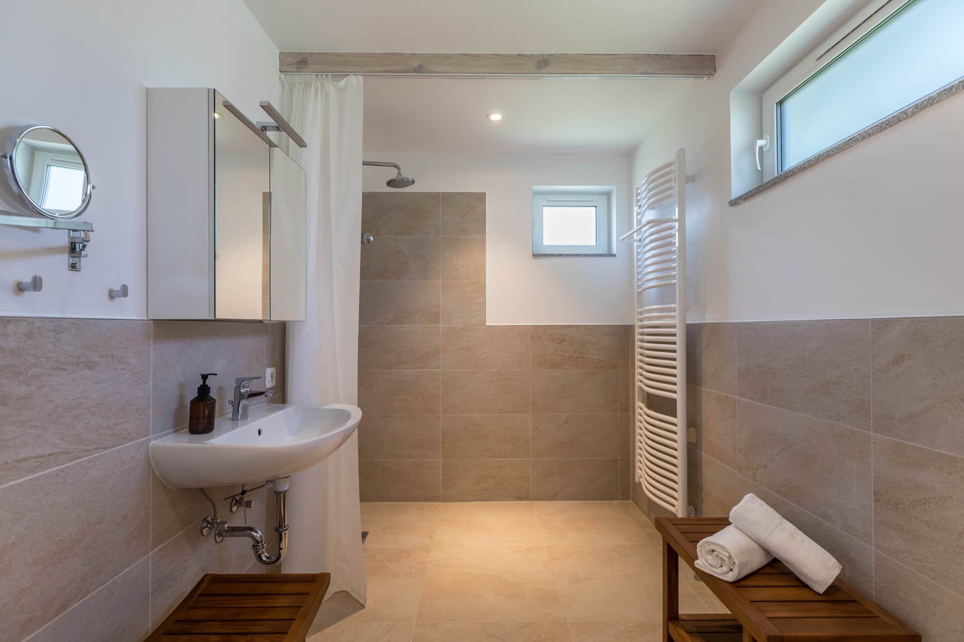 A bathroom with beige tiles: the basin under the mirrored cabinet, a shaving mirror beside it and the rain shower behind a pale curtain.