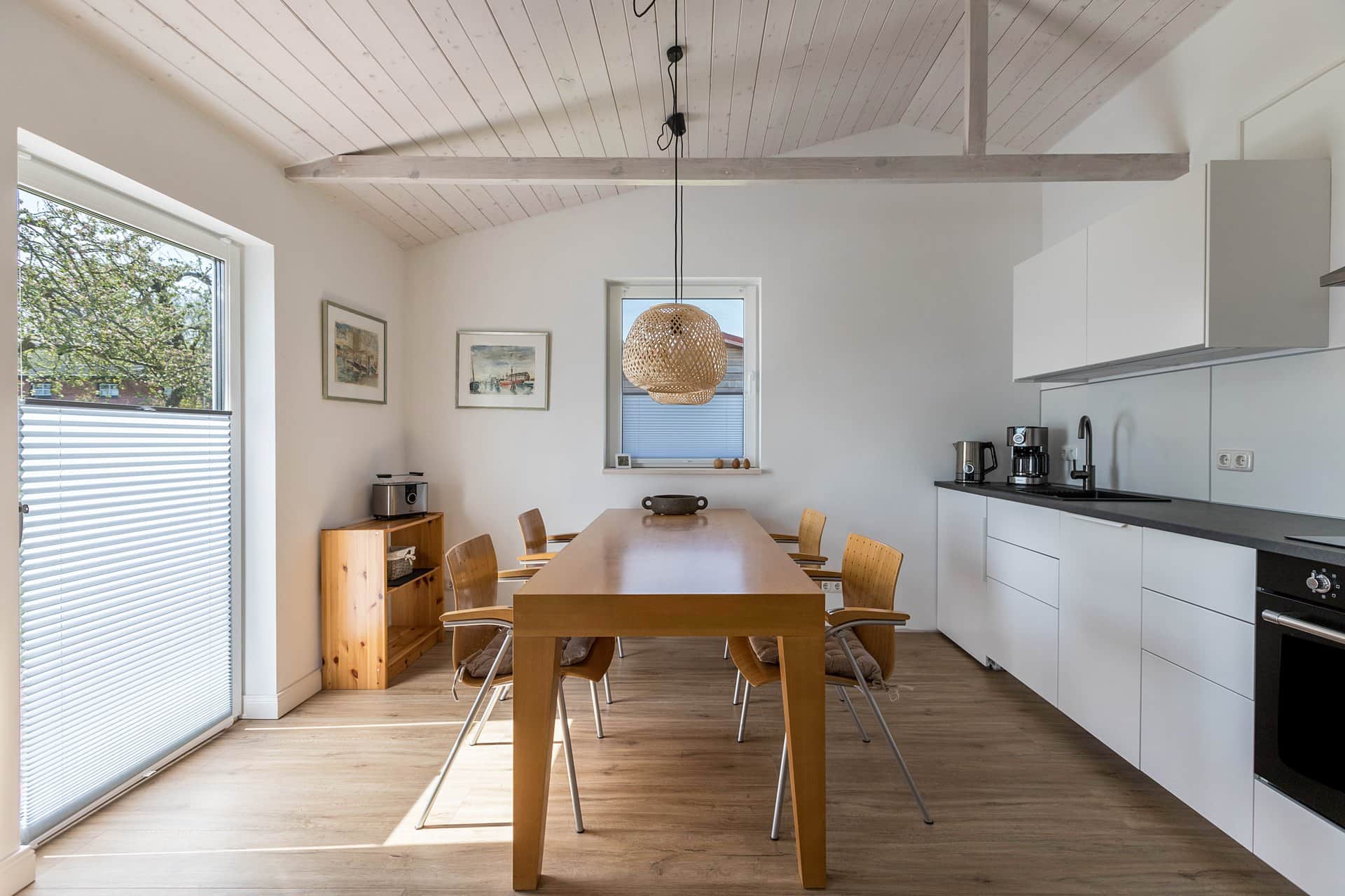 A dining table with six chairs beside the white kitchen units with their black worktop; a floor-to-ceiling door on the left and framed pictures on the wall.