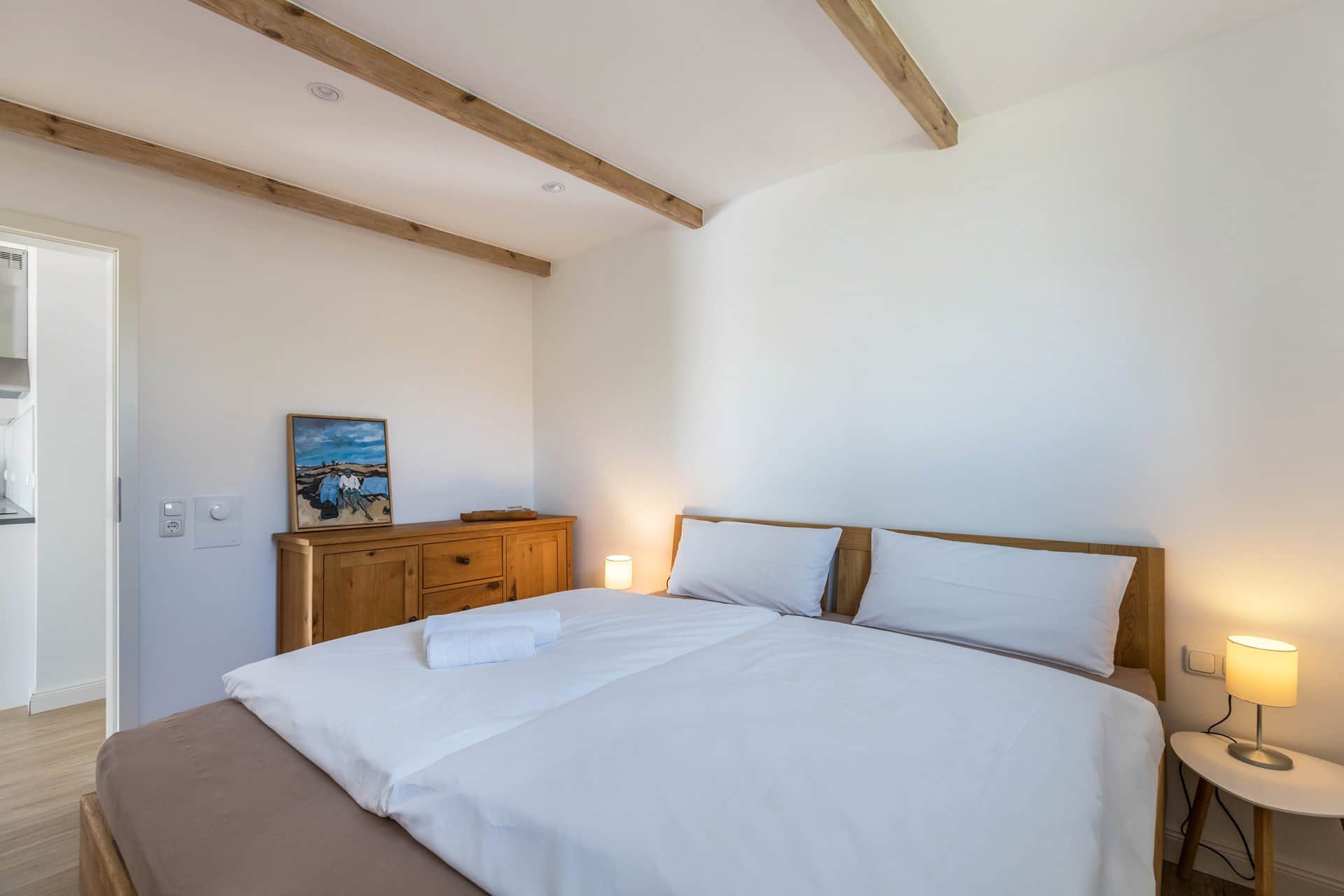 A double bed with white linen between two bedside lamps, with an old wooden chest of drawers and a picture on it to the left.