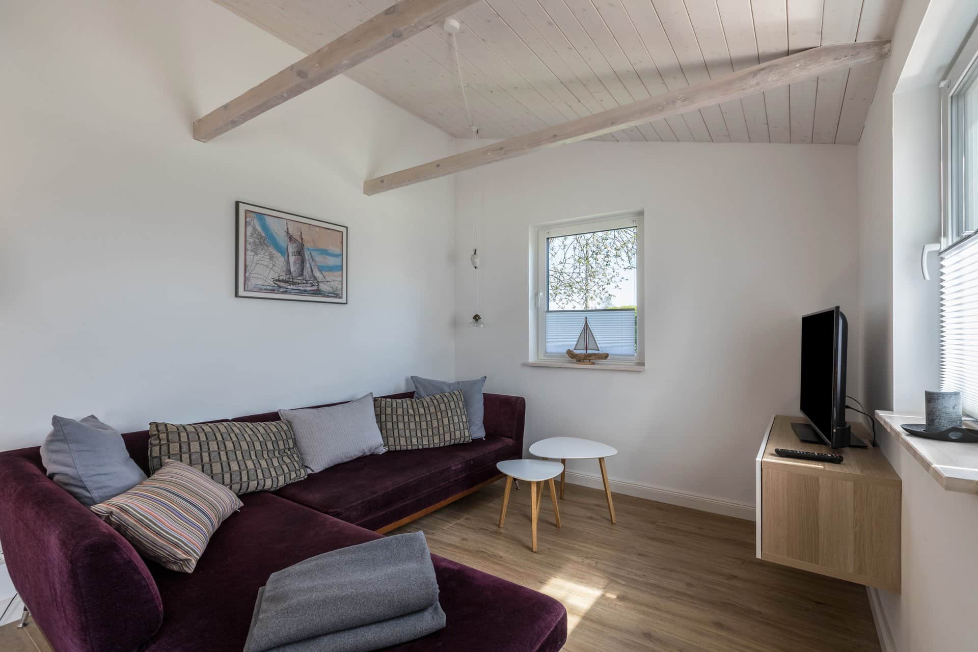 The purple corner sofa with patterned cushions and a folded blanket, two round side tables beside it and the television on the sideboard to the right.