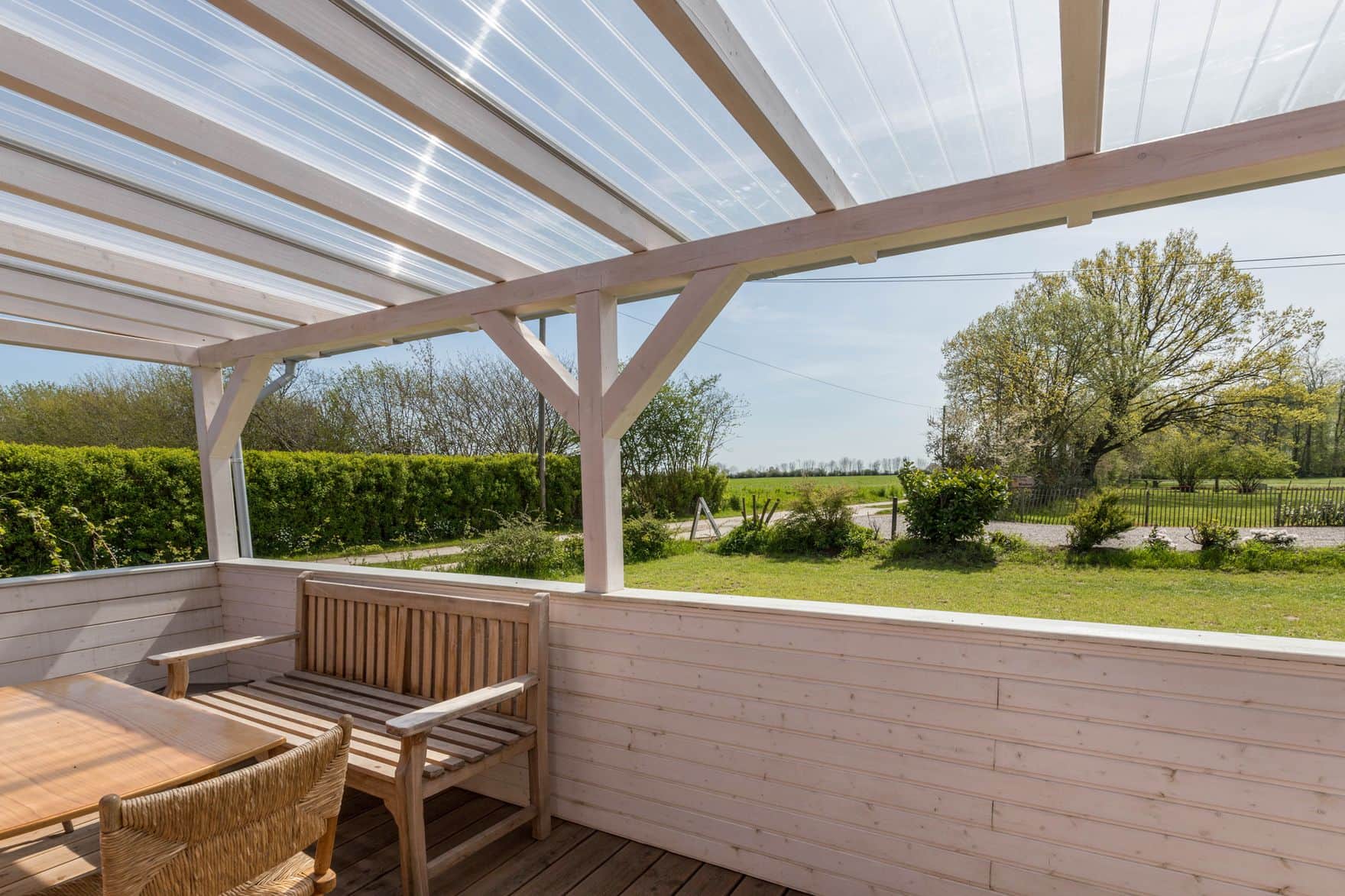 The covered veranda under translucent roof panels: a wooden bench and a wicker chair at the table, with the hedge, a field and the sky beyond.